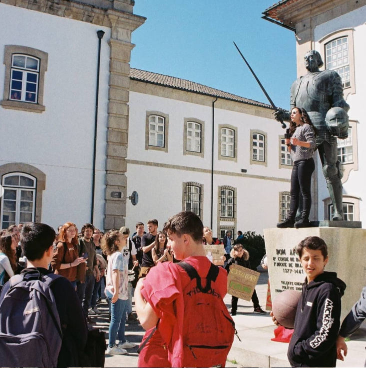 students gather around a statue monument where a young person is standing on top of the statue giving a speech